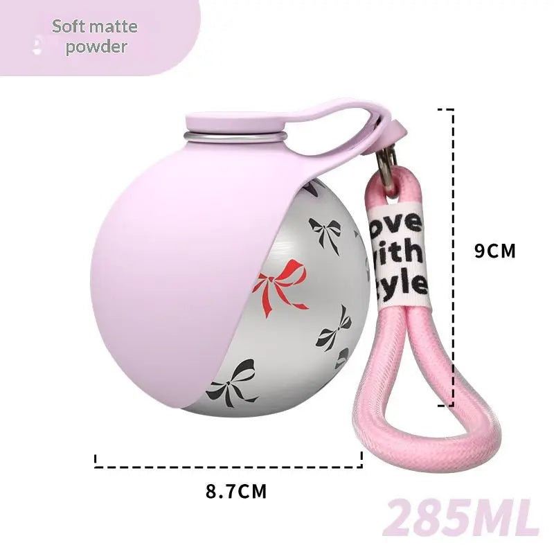 A 285ML matte powder bottle with a light pink silicone sleeve and a pink carrying loop. The bottle body is metallic silver with black bow patterns and a red bow accent.